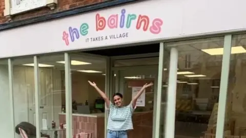 Henna Karim-Sayer Henna, in a green and white striped top, proudly poses in front of her shop sign with her arms aloft in celebration