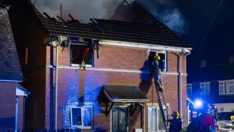A house with a badly fire-damaged roof and a fire officer climbing up to the window on a ladder. 
One of the top windows is badly damaged and black with no window panes.
There is smoke in the air and more fire officers in front of the house. 