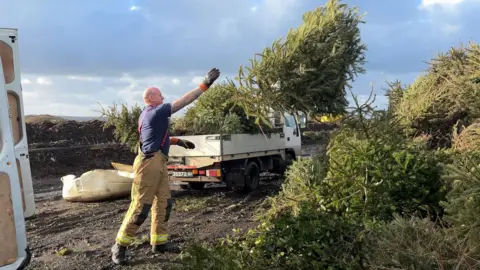 BBC Firefighter Charlie Roger in his uniform, throwing a Christmas tree on to a larger pile of trees. 