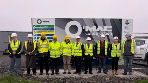 Ten representatives from SSEN Transmission and Hebridean Housing Partnership in hi-vis jackets and helmets, standing in front of a black and white construction billboard. Grey skies behind. Fencing, grey portacabin and white construction vehicle in the background. 