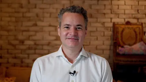 A man with grey hair and a white shirt looking at the camera. He is smiling and sat in front of a light brown brick wall. There is a small black microphone clipped to his shirt
