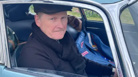 William Duncan is sitting inside the driver's seat of a blue vintage car. He's wearing a flat cap and has a scarf and jumper on. A leather driver's jacket is sitting on the passenger seat.