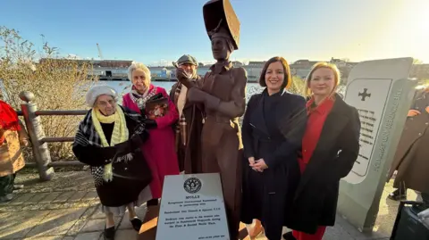 A group of four women and one man stand around a 6ft steel sculpture called Molly on the banks of the River Wear in Sunderland. In front of the sculpture is a plaque with more details about the artwork.