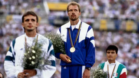 Getty Images Andrey Perlov on the podium at the Olympic Stadium in Barcelona in 1992, wearing a blue and white tracksuit with a gold medal round his neck, holding a bunch of flowers - on his left and right are the competitors who came second and third in the race