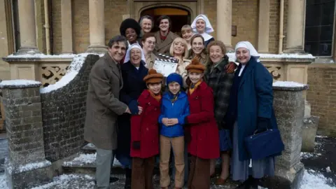 Cast members of Call the Midwife dressed in period clothing stand on a set of snowy steps holding a clapper board