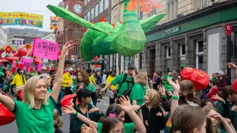 A crowd of children raising their hands up to the sky as they walk through a street, with a large green puppet over their heads