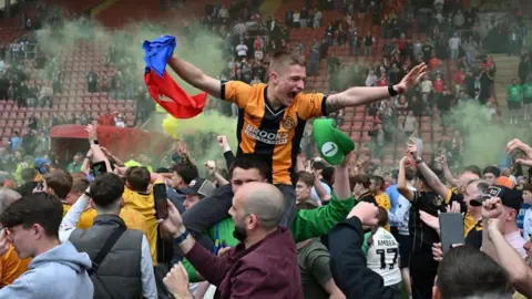 Fans celebrating inside the stadium - with one fan in a yellow and black jersey sitting on the shoulder of another fan. 