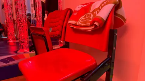 Image of two red fold-down stadium seats, with a red throws on the backs of each seat. In the background can be seen three tall water filled tubes with bubbles and some people playing behind them.