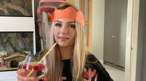 The Stroke Association Jo Dayton with long blonde hair wearing an orange paper crown and a dark jumper with orange sparkly antlers on. She is holding a gin glass with raspberries and a yellow and white straw in. Multiple painting are hung on the wall behind her and there is an open wooden door. 