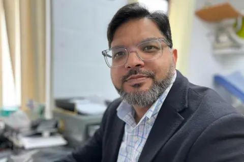 Dr Amir is sat at his desk in his consultation room, looking at the camera with a neutral expression. He has dark hair and a beard and is wearing a checked shirt and dark blazer, with light rimmed glasses.
Behind him, out of focus, are office supplies on the desk.