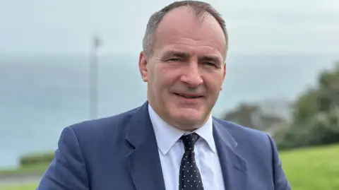 Alfred Cannan, who has short dark hair and it wearing a blue suit and tie with a white shirt. He has a headland and the sea behind him.