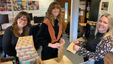 Three women, one with glasses, one with a fringe and one with blonde hair, are standing inside an office wrapping Christmas presents.