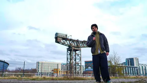 A large industrial crane on an urban riverside area, a person holding a microphone stands in the foreground.