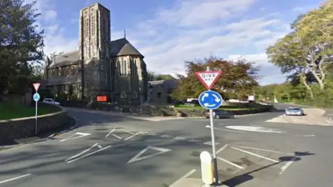 A roundabout with the Braddan Church building on one corner and a Give Way sign in the foreground on a clear day. There are trees on the right hand side and the sky above is blue with some wispy white clouds.