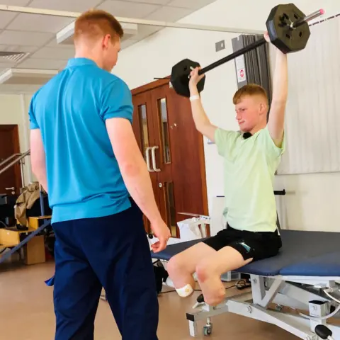 Adam Golebiewski, teenager with both legs removed below the knee, sitting on a hospital bed in shorts and a green tee shirt and lifting weights above his, with orthopaedic rehabilitation physiotherapist Owen Cairns, in blue trousers and a blue tee shirt, standing instructing.