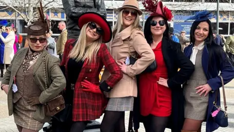 Five young women pose with their hands on their hips side on to the camera. They are all wearing tweed or smart jackets and dresses with hats and fascinators.