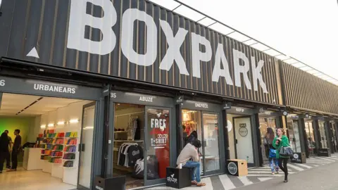 Getty Images We can see several containers are lined up next to each other with shops inside them. About them is a big sign saying BOXPARK, which is printed on corrugated metal.