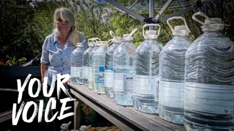 Genista Tanner stands beside an outdoor wooden table covered with large clear plastic containers filled with water. The containers are arranged in a line and dominate the foreground of the image. Behind them is a garden setting with trees, plants and outdoor furniture. The photograph illustrates her reliance on stored bottled water after the loss of a private household water supply.