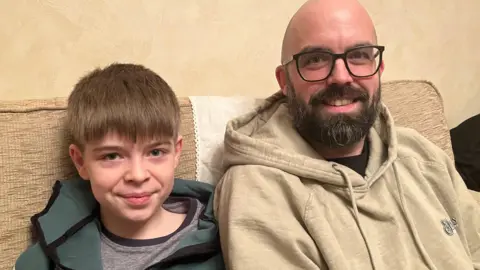 Theo wears a green top and is sat on a sofa up against his father to his right. He has short brown hair brushed forward, while his dad has a bald head, black beard and is wearing black-rimmed glasses.