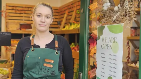 Carissa Patten, wearing a black top and green apron, stands in the open door of a greengrocers shop