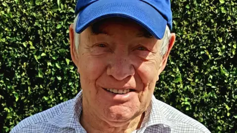 Lynsey Marsden An elderly man smiles at the camera. He has a chequered shirt and royal blue Puma cap on. Behind him is a hedgerow, and it appears to be sunny.