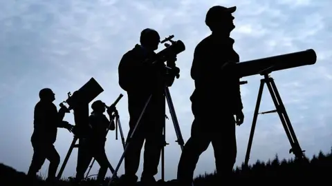 Getty Images Silhouettes of astronomers setting up telescopes and cameras ahead of a solar eclipse. The sky is clouded over.