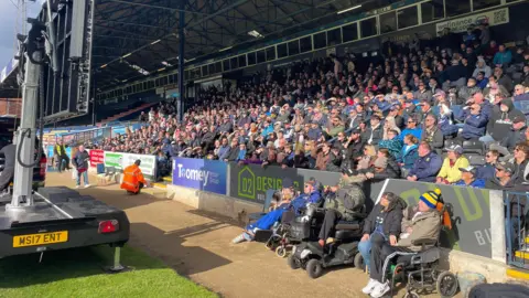 Stephen Huntley/BBC A crowd fill seats in a section of stadium. People are all sat in their seats, looking at a big screen mounted on a trailer.