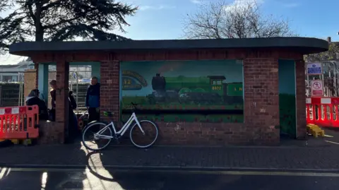Sheringham bus shelter. It is a brick building with a flat roof built in an art deco style. There is a mural of a train on the inside. There is a bicycle leaning against the front of the shelter. There are three people inside. They are gathered on the left hand side. Behind and beside the shelter there is wire fencing and orange-and-white barriers.