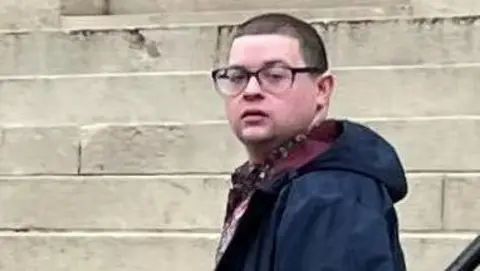 Ieuan Crump wearing a long navy coat and dark glasses, standing in front of steps