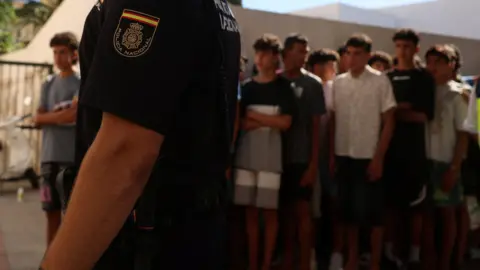 A police officer stands guard as migrant children queue outside a police station in Ceuta to be registered following mass crossings by migrants on foot and by sea from Morocco.