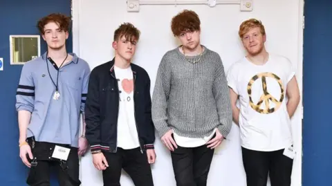 Four men stand side by side in front of a white wall outside recording studios. They are all looking at the camera.