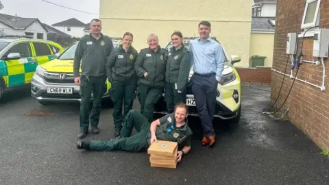 Jordan Harries A group of NHS staff stood with Jordan in front of emergency vehicles. There is an NHS worker lying on the ground in front of the others holding the pizza boxes that were donated.