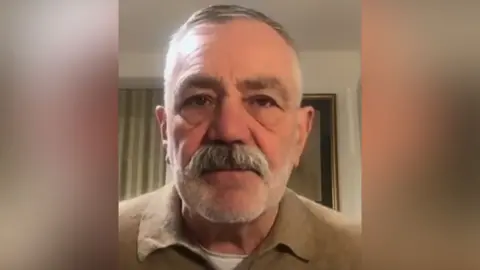 David Wilson looks into the camera. He has white stubble and a grey moustache. He is wearing a light brown jumper.