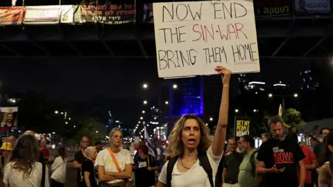 Reuters A woman in a white t-shirt stands in front of a crowd of demonstrators at night. She holds a placard that says 'now end the sin-war, bring them home'. 