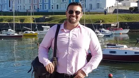 Andrew Fearon is photographed standing outside in front of a harbour, which has several boats in it. behind the harbour is a large patch of grass and colourful houses. He is wearing dark sunglasses, a pink shirt and has a bag to his left side. He is smiling and looking at the camera.