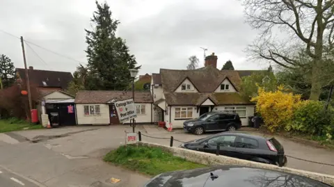 A street view image of a white-coloured village store and post office. Cars are parked outside the front of the store.