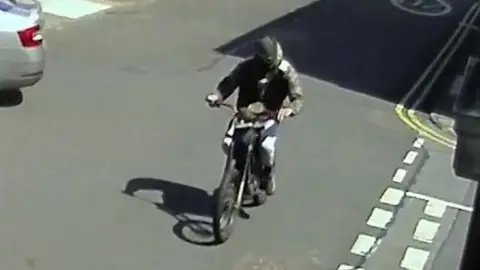Northumbria Police Still from CCTV looking down on to a road, along which Billy Stokoe is riding the electric motorbike. He is wearing grey trousers, a camouflage jacket and a black balaclava.