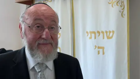 The Chief Rabbi wears a dark suit, white shirt, grey tie with a darker grey square pattern, and a kippah. He wears glasses and has a white beard.