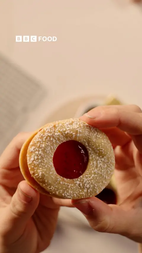 A pair of hands hold a round biscuit with red jam in the middle