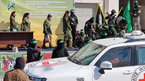 Getty Images The four female Israeli hostages begin walking off stage as Hamas fighters armed with guns and wearing black with their faces covered watch on. A car with the logo of the Red Cross on it is parked next to the stage