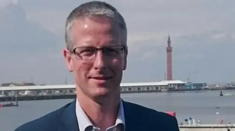 Rob Walsh with short-cropped grey hair. He is wearing dark-metal framed specs on his face, a navy blazer over a white and blue thin-striped shirt. He is standing on the dock with Grimsby's Dock Tower in the background and a row of one-storey buildings.