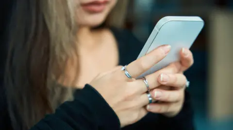 A close-up image of a young woman holding a smartphone, wearing silver rings on her fingers.