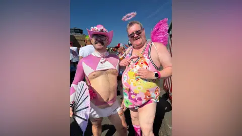 Two men in pink outfits. One has a fluffy pink halo, the other a neon pink cowboy hat.
