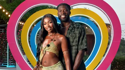 ITV/REX/Shutterstock Mimii and Josh, two contestants from Love Island, smiling and standing next to each while a heart-shaped backdrop is behind them in the colours of blue, yellow and pink