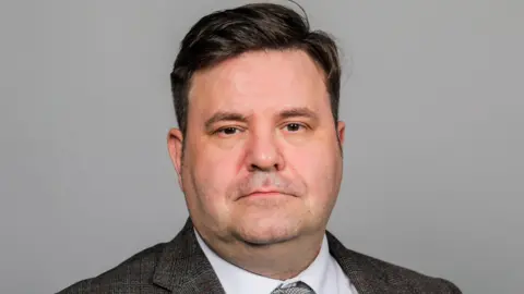 A head and shoulders shot of Alec Brown who has dark hair and is wearing a grey suit and tie.