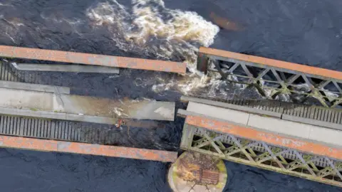 A collapsed iron girder bridge with a section between two stone piers breaking apart and falling into the water.