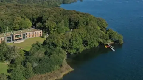 An aerial view of the YMCA Lakeside camp in Windermere, which is next to the lake. It has a modern building with a big box window next to it and is surrounded by trees. There is a jetty.