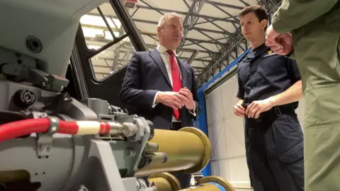 Luke Pollard speaks with crews next to a wildcat helicopter in a hangar, there are two men next to him
