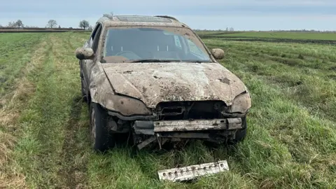 Mathew Latta A 4x4 car has been abandoned in a field. It is covered in mud and the front part of the vehicle is hanging off. 