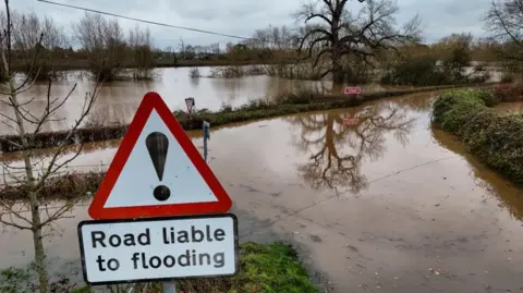 Widespread flooding near Upton-on-Severn, after Storm Bert. In the foreground, a warning sign advises that the road is liable to flooding and indeed, both the road and all surrounding fields are submerged under brown, muddy water.